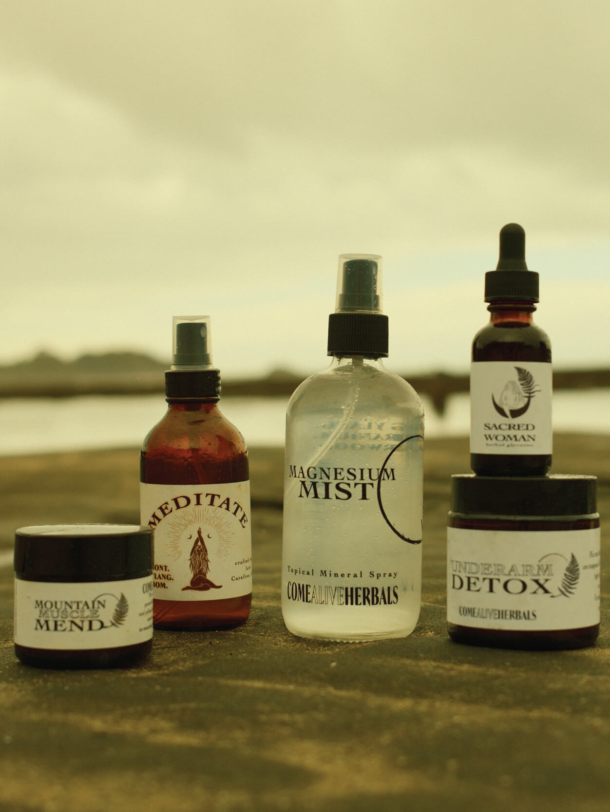 five products from left to right; mountain muscle mend, meditate mist, magnesium mist, sacred woman (sitting on top of), underarm detox sitting on a sandy beach with blurry waters edge background.