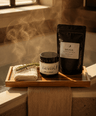 A spa-style detox product set displayed on a wooden tray in a warm, steam-filled bathroom setting. The set includes a black stand-up pouch labeled 'DETOX MELANINIC PINK', a matching black jar labeled 'DETOX', a folded white towel, and a sprig of greenery.