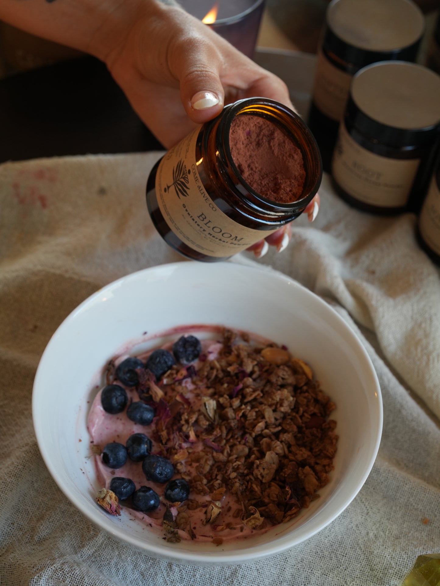 A hand holds an amber glass jar of Bloom herbal powder over a white bowl containing yogurt, blueberries, and granola, set on a beige linen cloth with other jars blurred in the background.