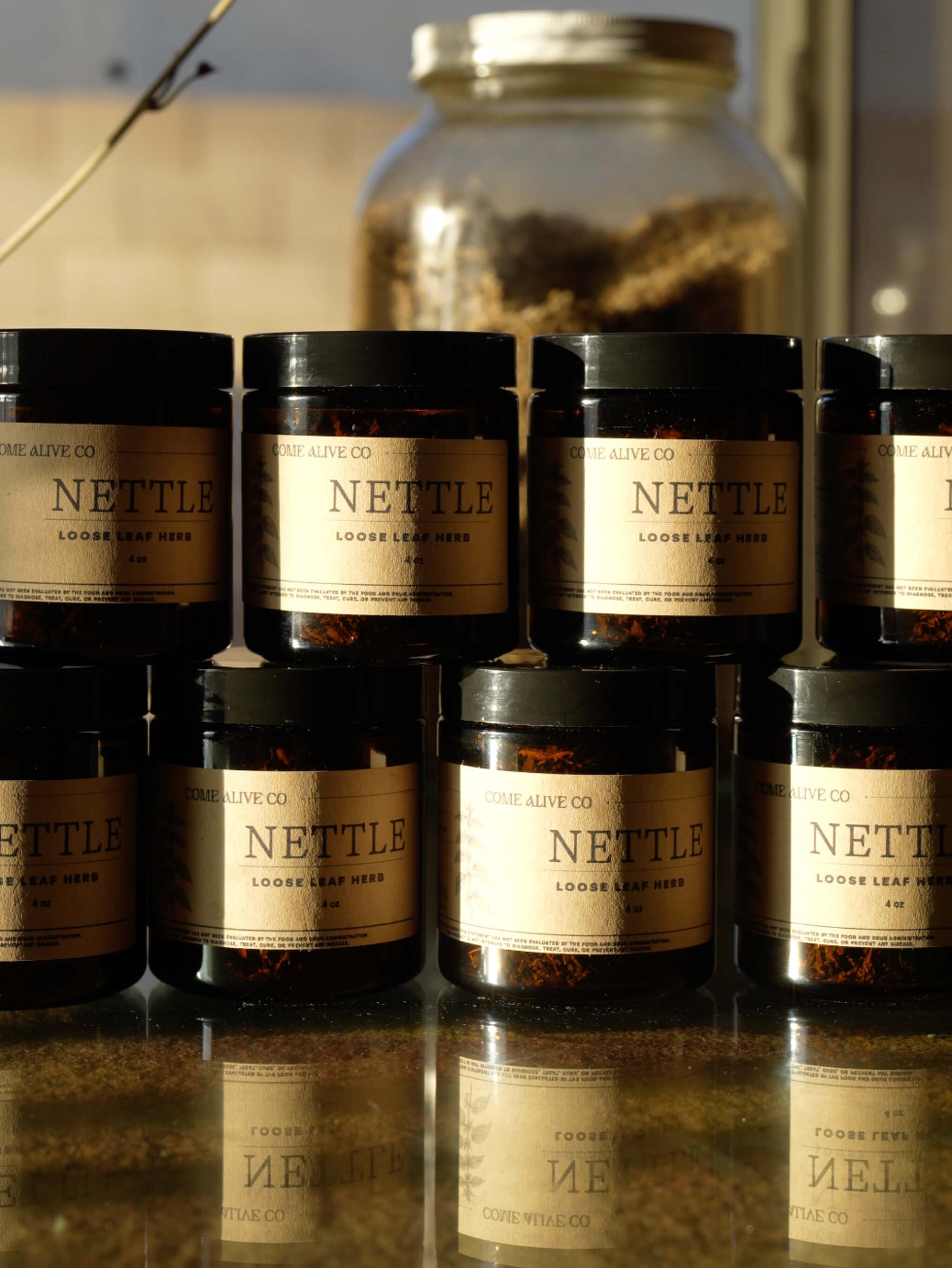 A group of amber glass jars filled with loose leaf herbs, labeled 'NETTLE' by Come ALiveCO.', arranged on a reflective surface with a larger jar blurred in the background.