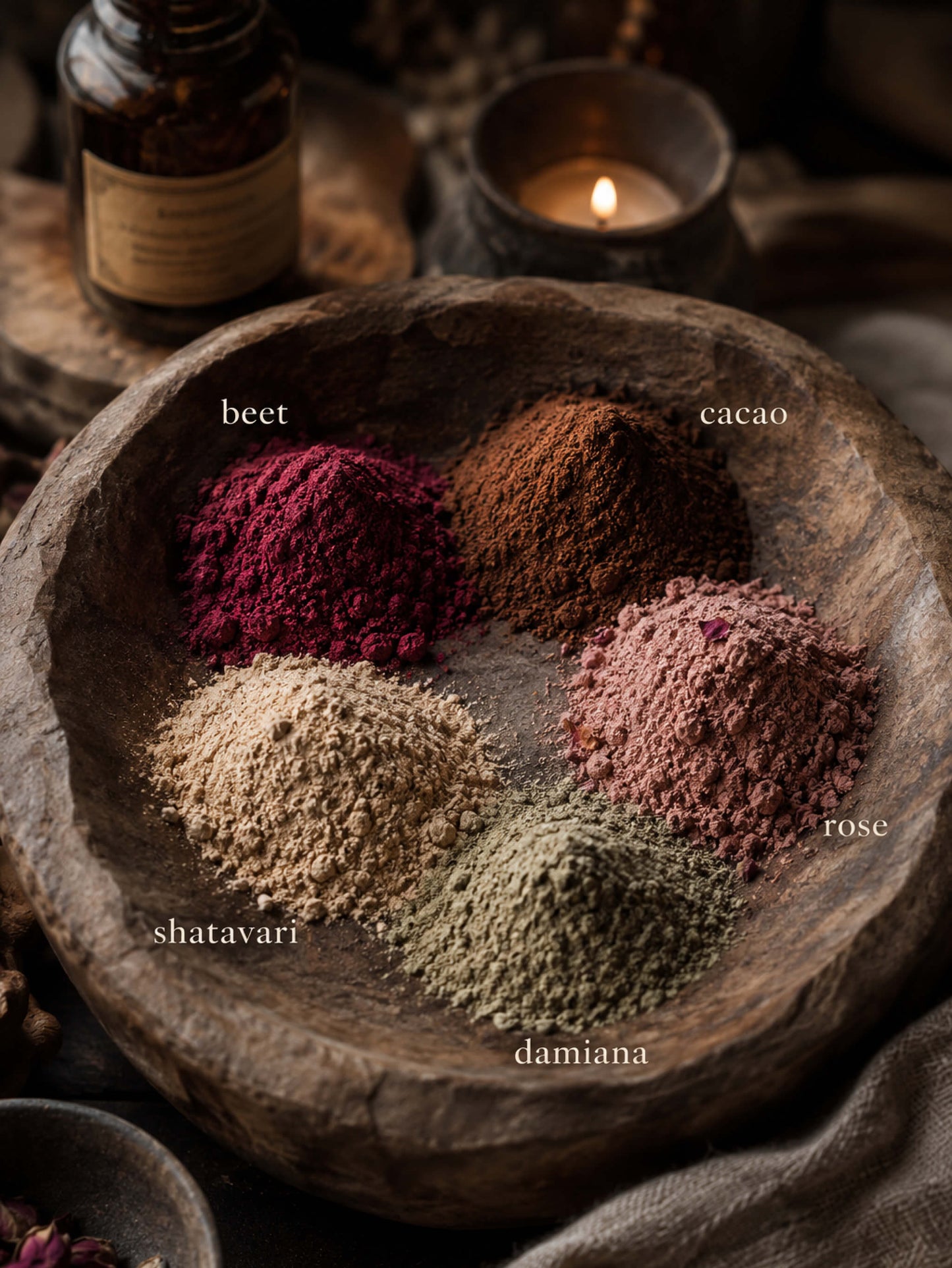 Five loose herbal powder piles arranged in a rustic wooden bowl, labeled beet, cacao, rose, shatavari, and damiana, with a bottle and candle blurred in the background.