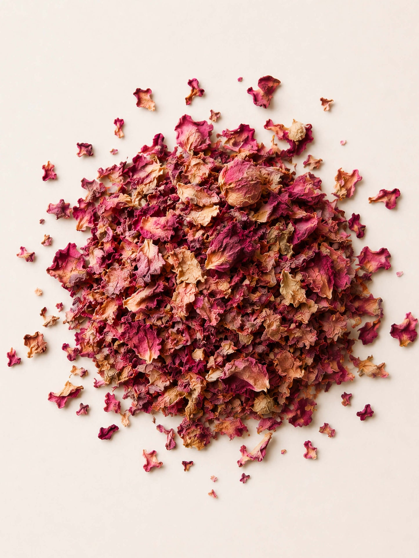 A pile of dried rose petals or flower petals in shades of pink, burgundy, and tan, scattered on a plain light beige background.