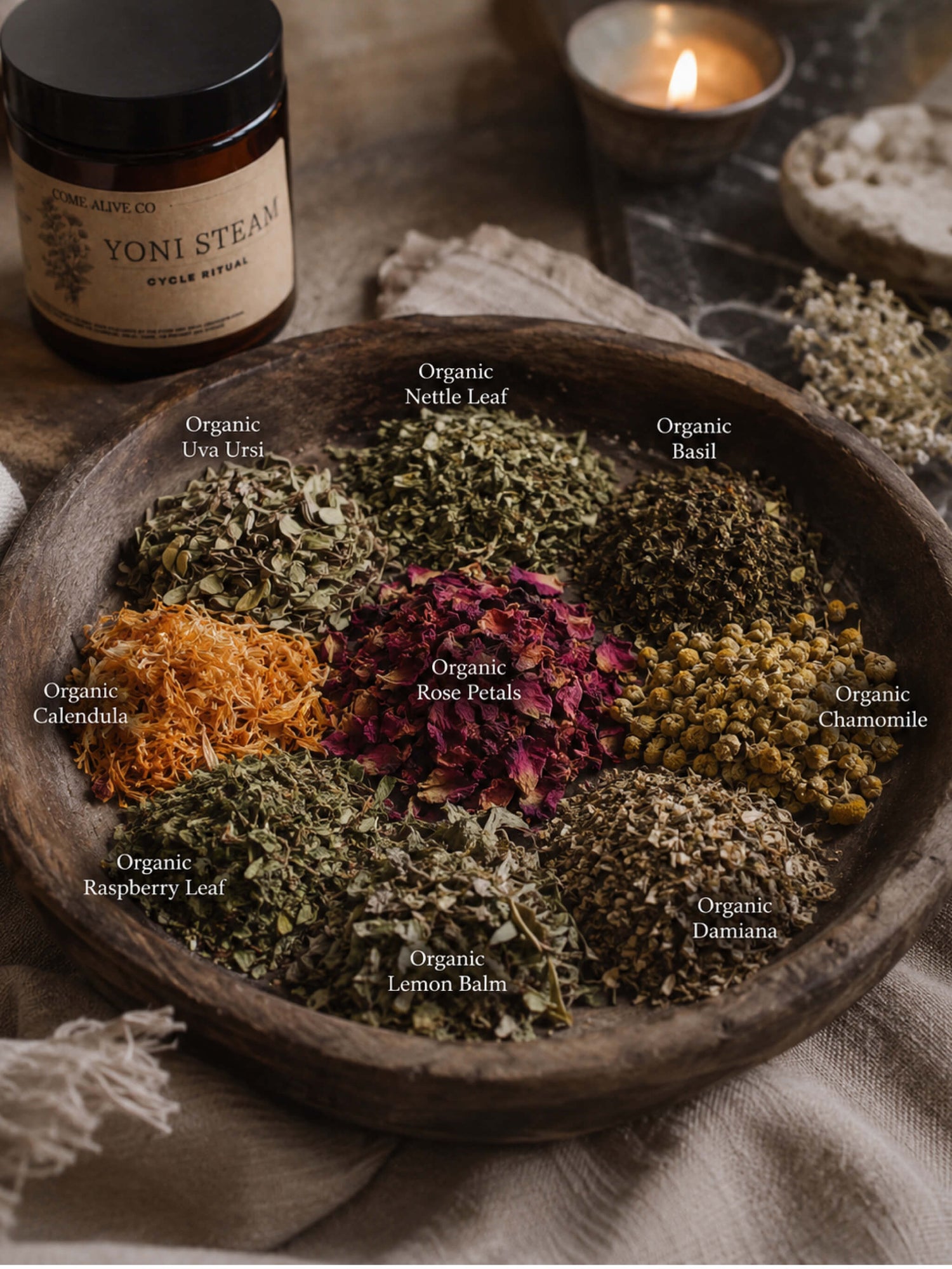 A wooden bowl filled with various dried botanical teas and herbs, arranged in a circular pattern with labels identifying each ingredient. In the background, a jar of Yoni Steam herbal tea and a lit candle are visible on a rustic fabric surface.