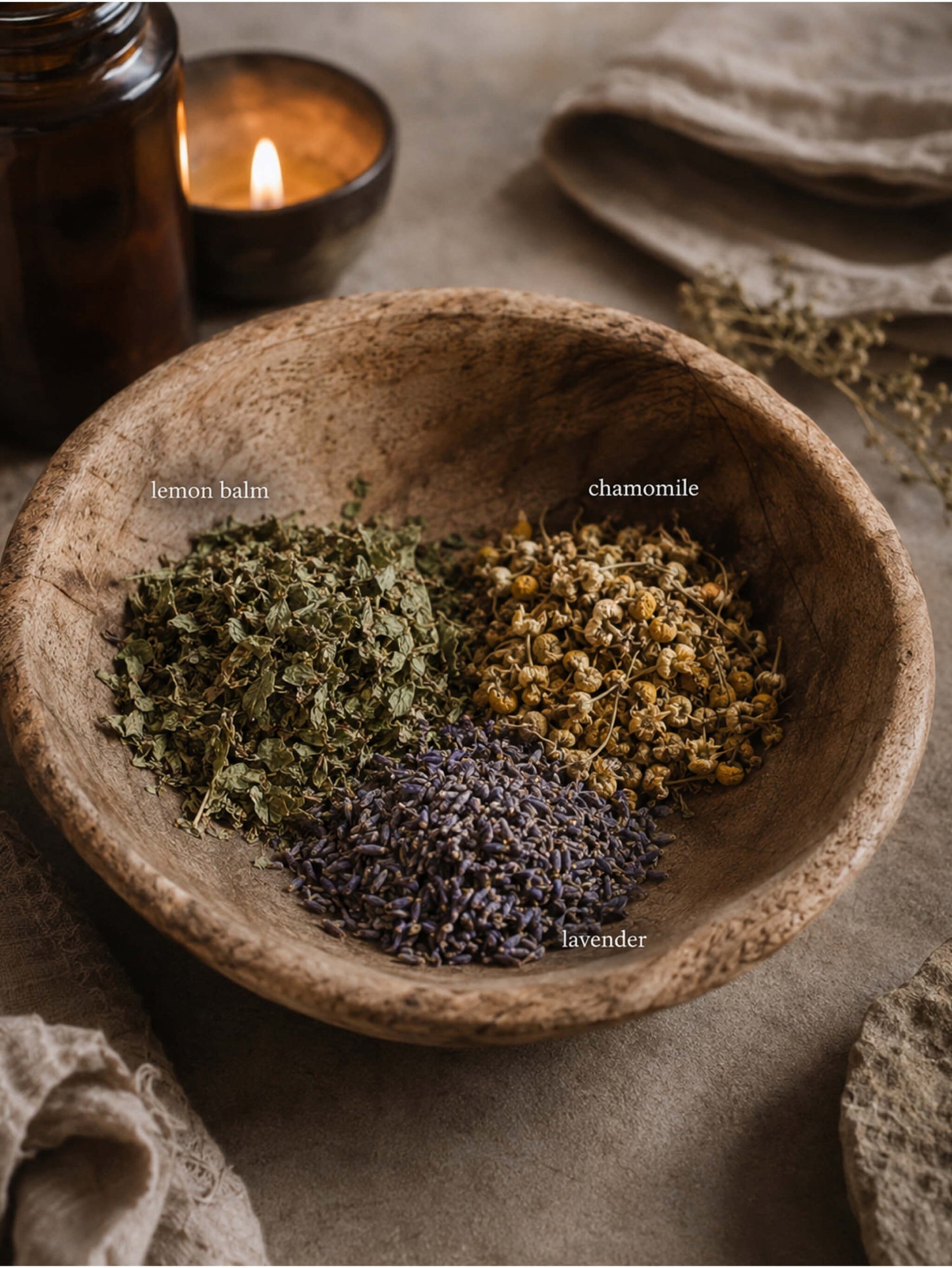 A rustic wooden bowl filled with dried herbal ingredients labeled lemon balm, chamomile, and lavender, styled on a linen surface with a lit candle and amber bottle in the background.