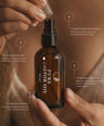 A person holds an amber glass spray bottle labeled PURE CASTOR OIL while applying a clear cap to the nozzle. The background is a warm brown skin tone, and the image includes four white text callouts with bullet points about benefits.