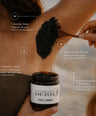 A person applies a thick black detox scrub from a jar labeled 'UNDERARM DETOX COMBAT HYPERHIDROSIS' onto their underarm with a wooden spoon. The image includes text callouts describing benefits and a bottom banner with product attributes like '100% natural', 'Vegan', and 'No aluminum'.
