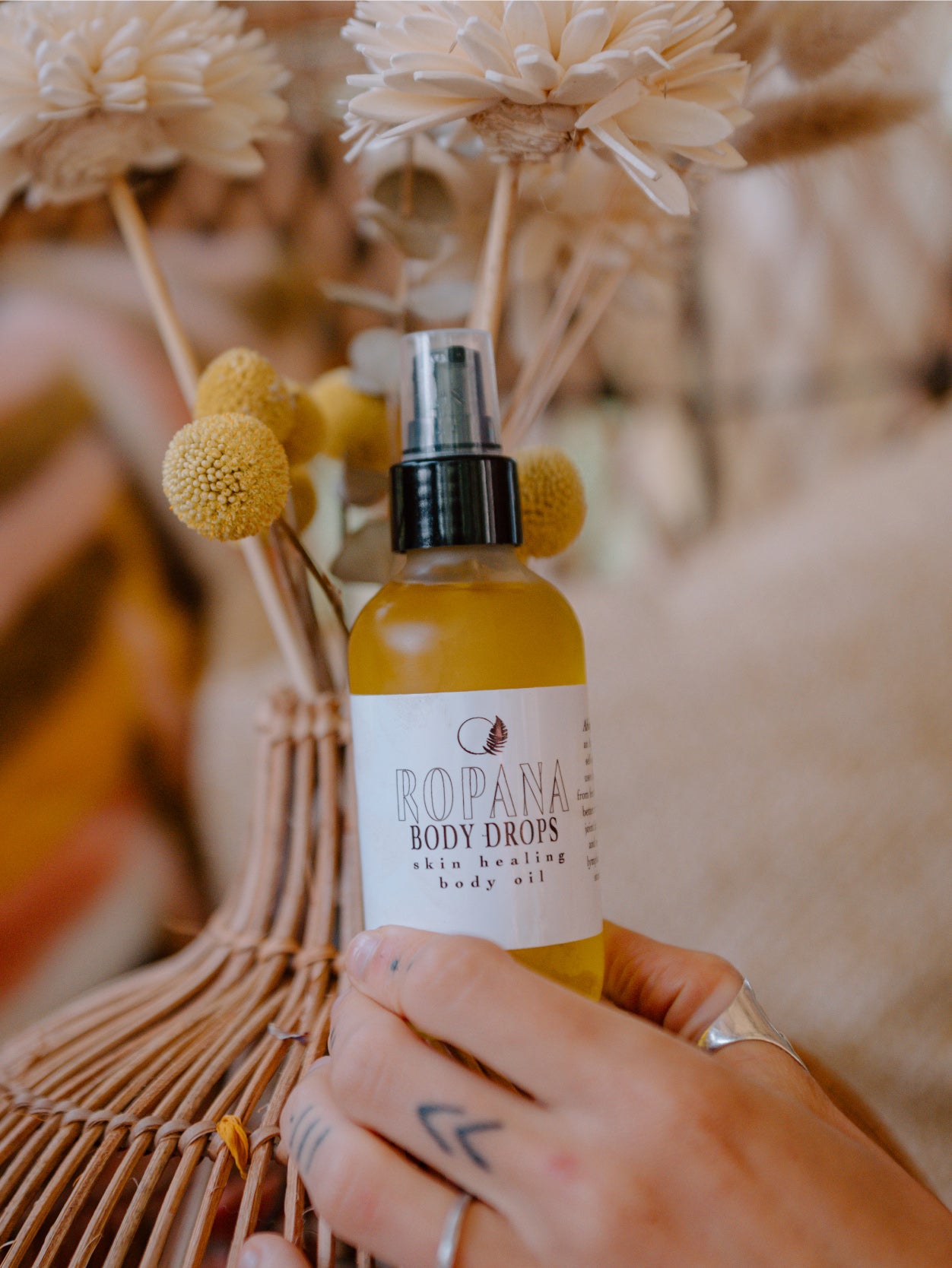 Frosted glass bottle with black spray lid labeled ropana body drops with a floral arrangement backdrop. 