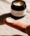 A close-up of a finger holding a dollop of white eye cream in front of an open dark amber jar labeled 'TALLOW EYE CREAM' on a beige fabric background.