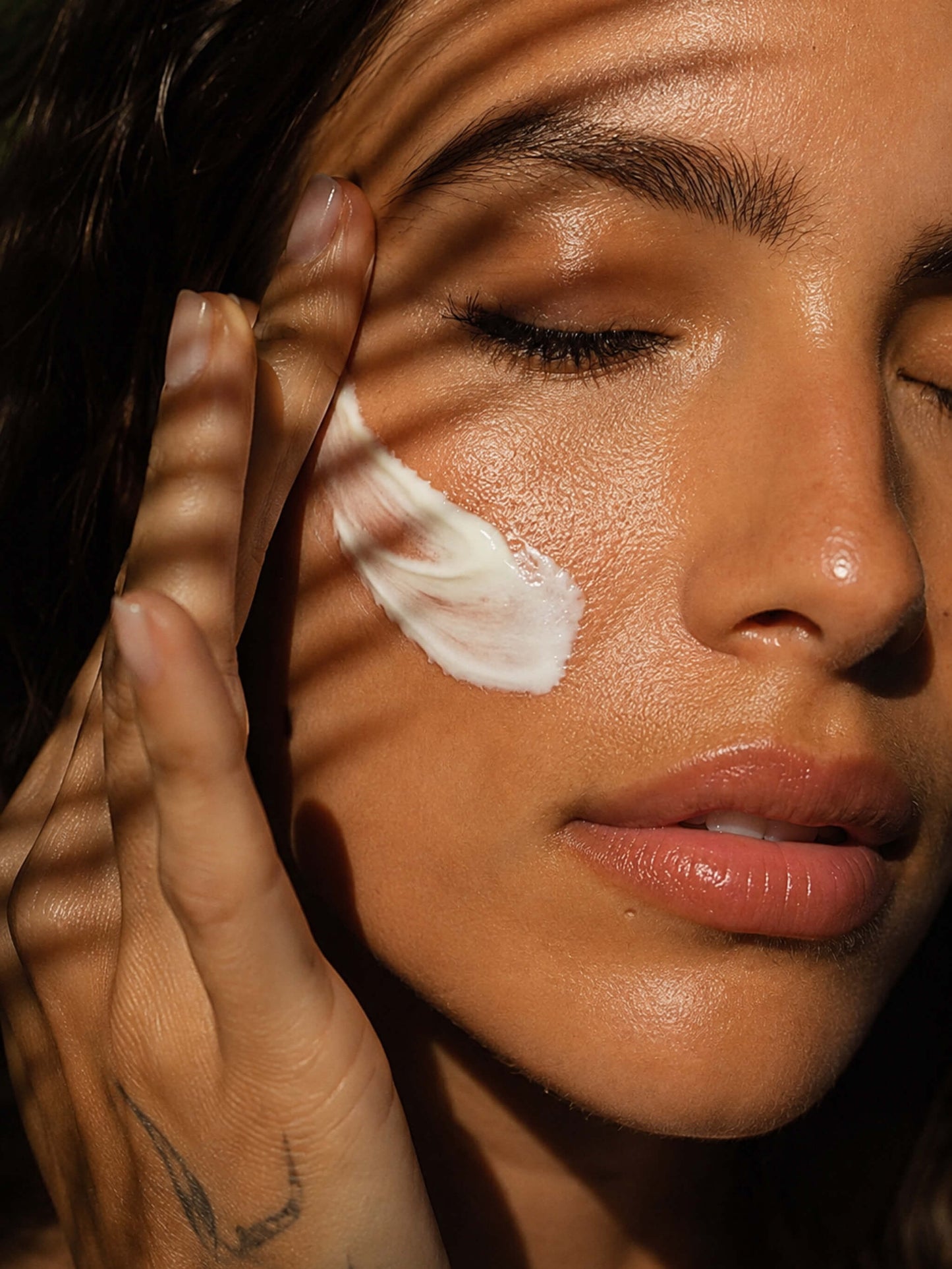 Close-up of a woman's face with closed eyes, applying or showing a white cream swatch on her cheek, with warm sunlight and leaf shadows cast across her face.