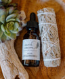 A glass dropper bottle labeled 'Nasya Drops' with a white label and green leaf design, next to a bundle of dried herbs, all placed on a wooden surface. Ayurvedic Nasya Drops for sinus support, breath clarity, and nervous system calm