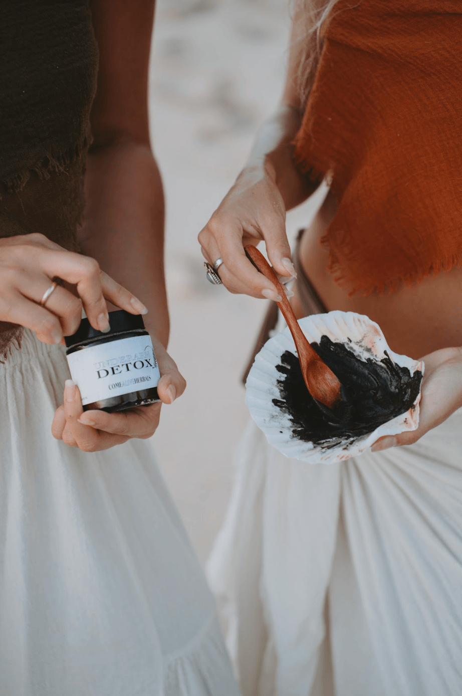 one woman hands holding amber glass jar labeled underarm detox while other woman's hands holding seashell with underarm detox paste in shell being mixed by wooden spoon with white background. 