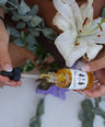 woman hand holding black glass dropper lid with yellow liquid while other hand holds clear glass bottle labeled yoni oil with floral background. 